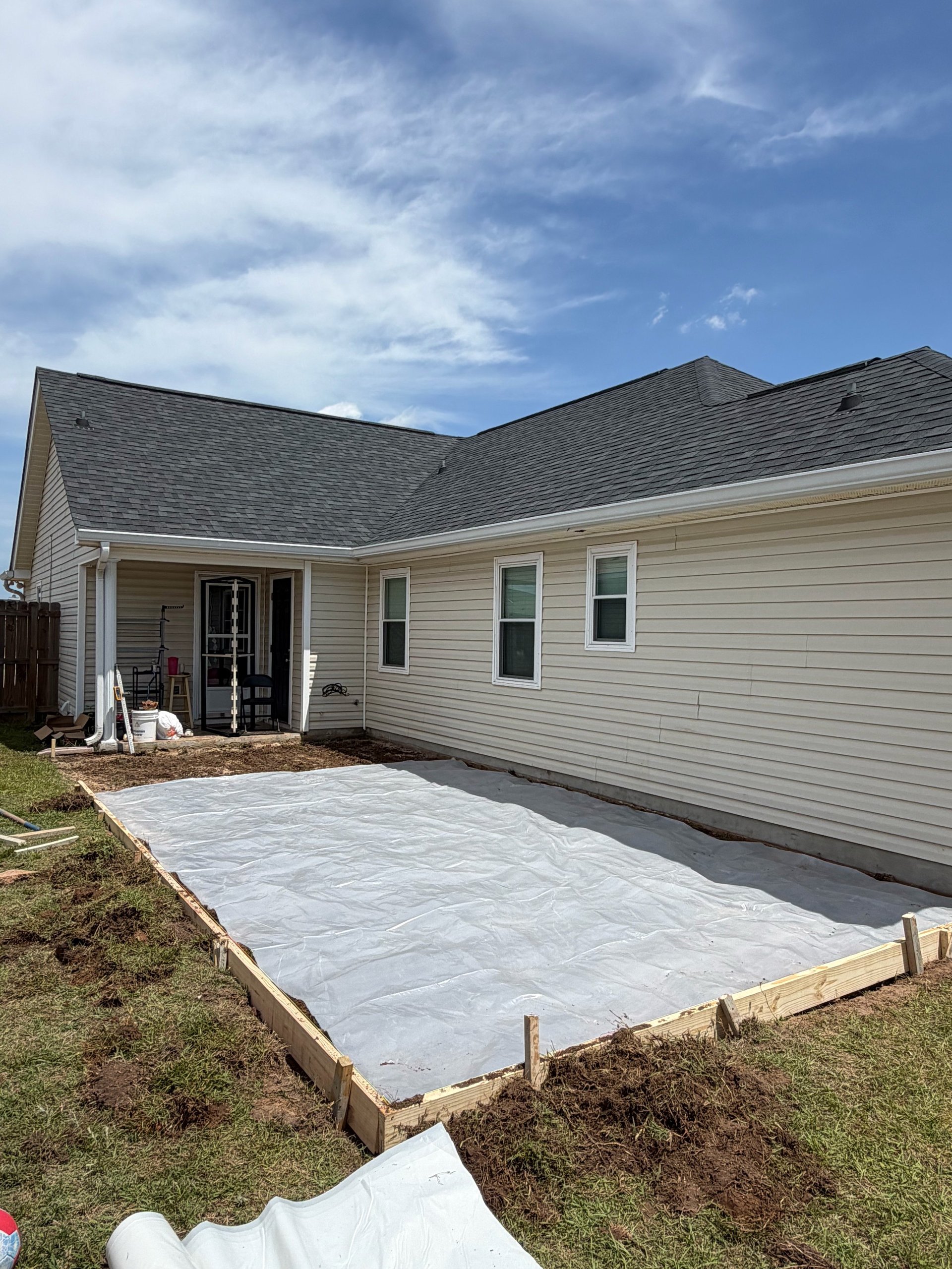 Single-story beige house with dark roof and back patio under construction, white foundation visible on yard under cloudy sky
