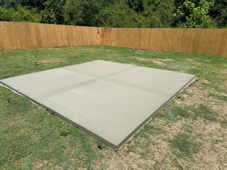 Gray concrete patio pad in backyard surrounded by green grass and wooden fence with trees in background
