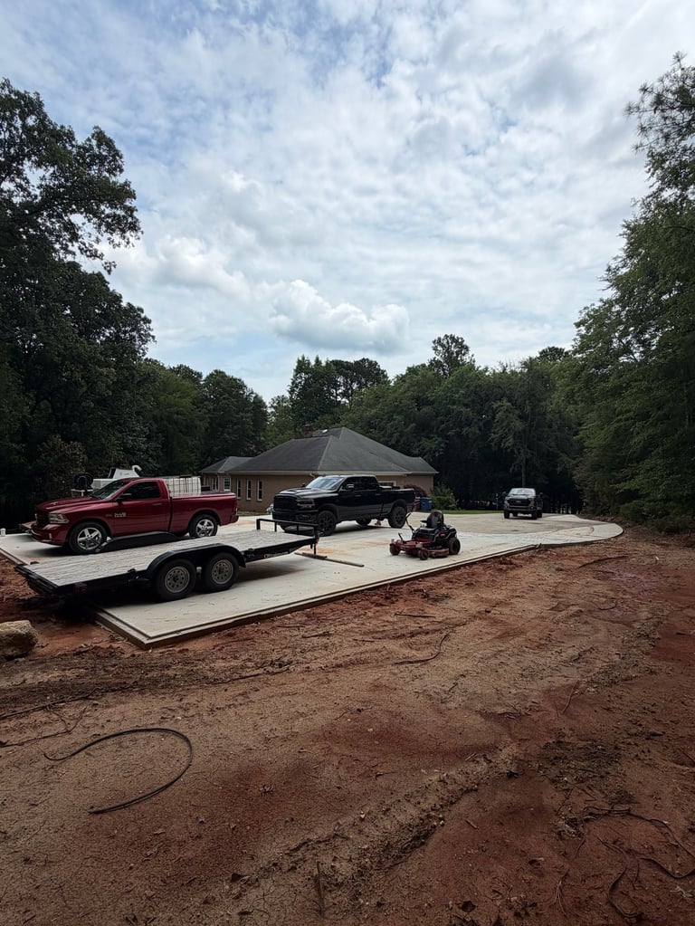 A concrete pad with a red truck on a trailer, black SUV, and red mower, set in a wooded area with a metal-roof building in background