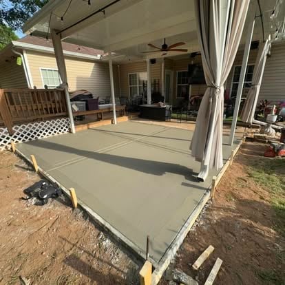 Newly poured concrete patio under construction with pergola and white curtains, attached to residential home with white siding.